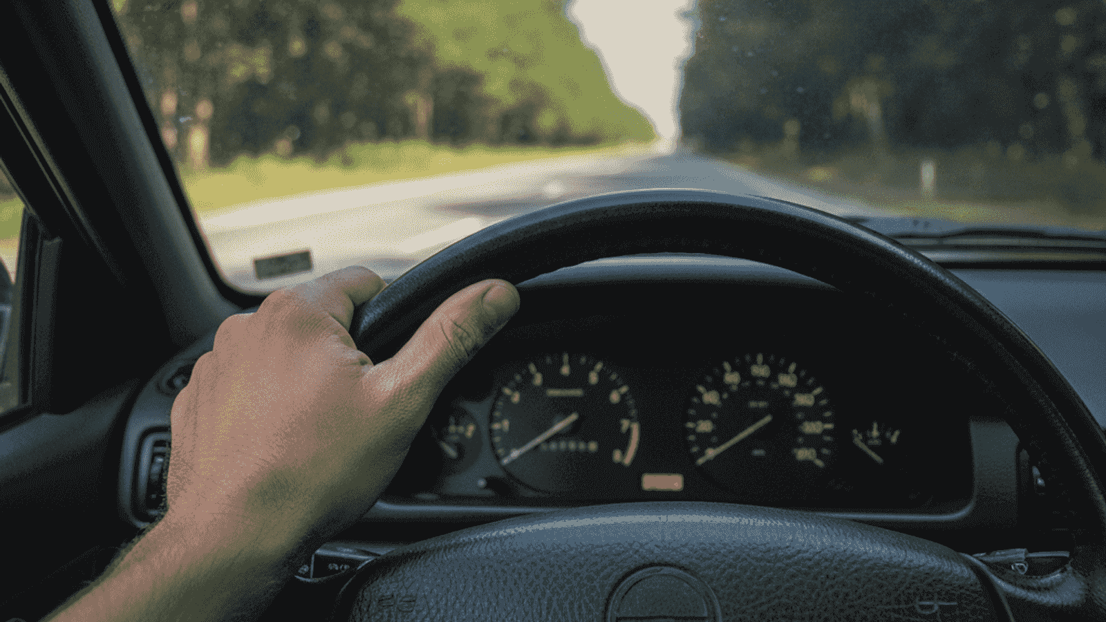 Driver's hand on the steering wheel of a European car with the dashboard showing low RPM during calm driving on a two-lane road