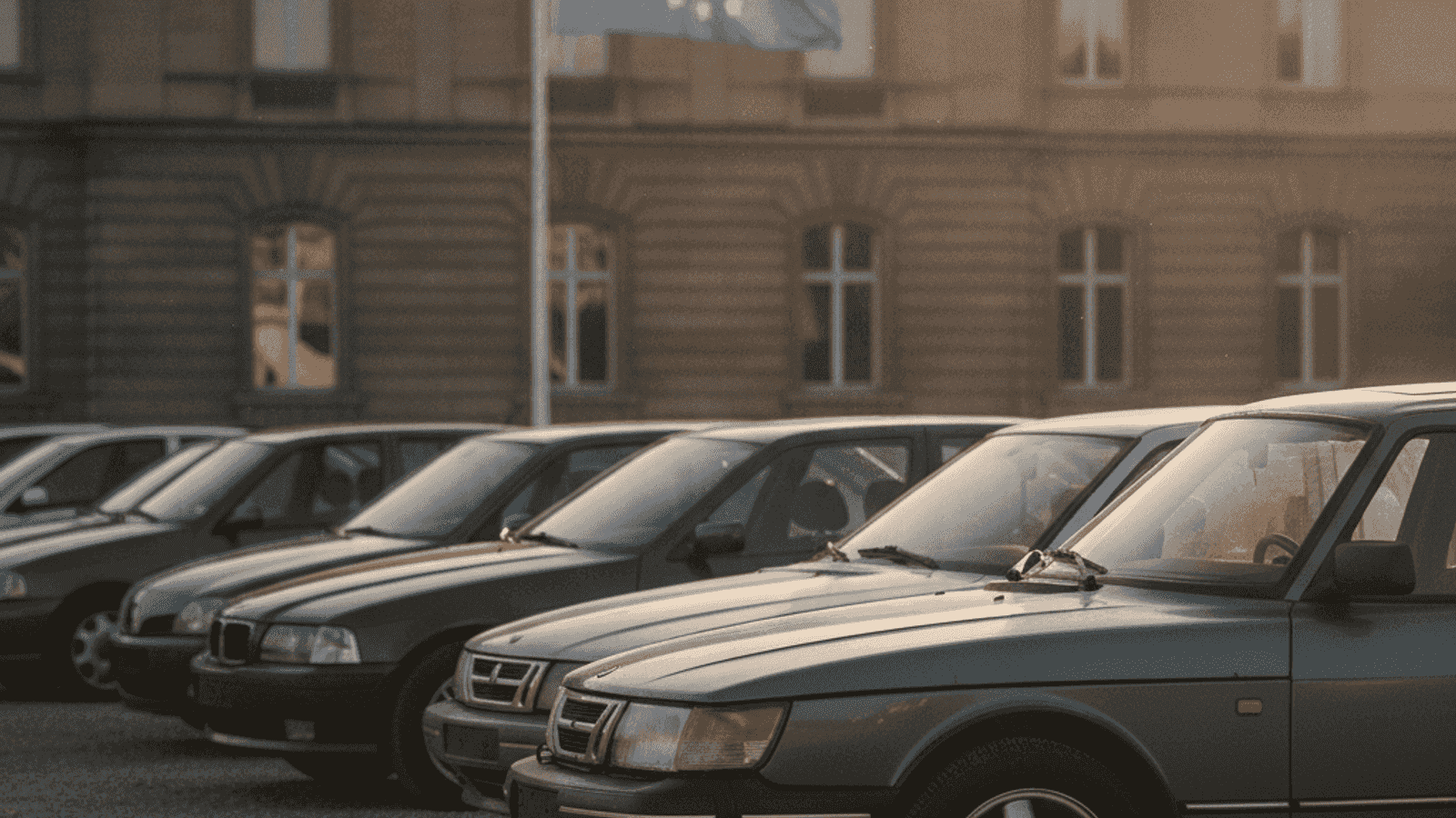Red starijih evropskih automobila na otvorenom parkingu sa EU zastavom u pozadini, toplo popodnevno svjetlo