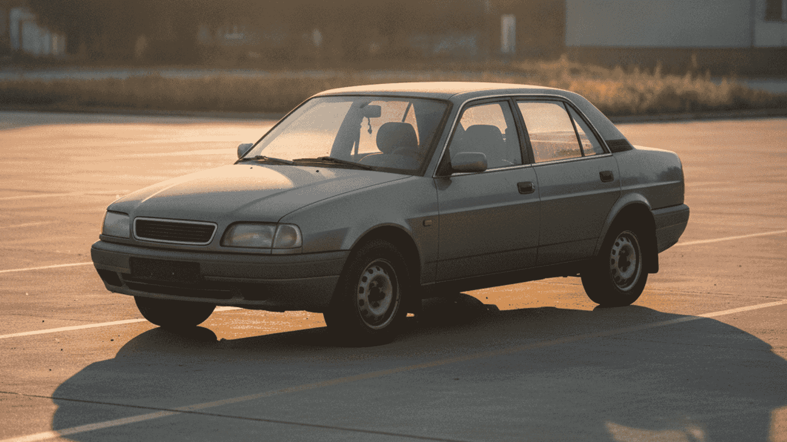 Stariji evropski sedan parkiran na praznom parkingu, prašnjav, sa blago spuštenim gumama i dugom sjenom od popodnevnog sunca