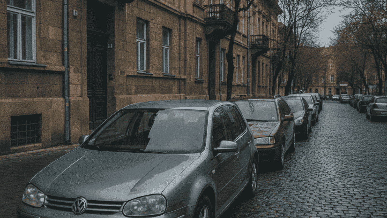 Ряд типичных европейских подержанных дизельных автомобилей, припаркованных на улице боснийского города, машины возрастом 10-15 лет под облачным небом