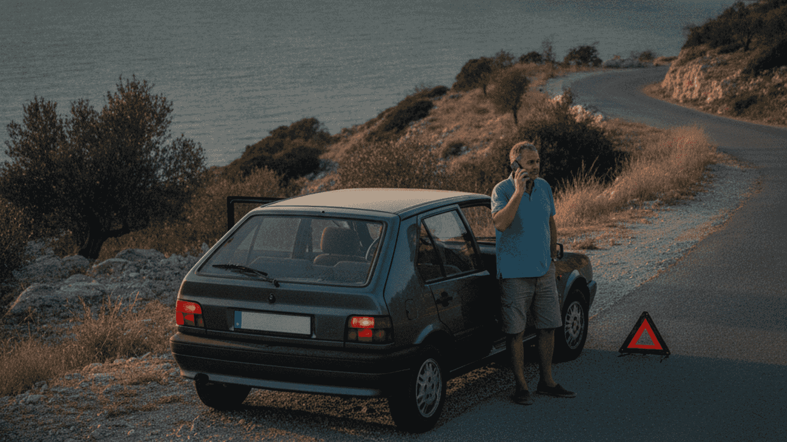 Car stopped on the shoulder of a coastal road with hazard lights and warning triangle on, driver talking on phone in afternoon light