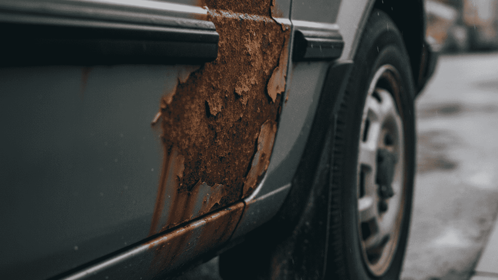 Close-up of rust on the lower sill and wheel arch of an older European hatchback with peeling paint and orange-brown corrosion on grey sheet metal